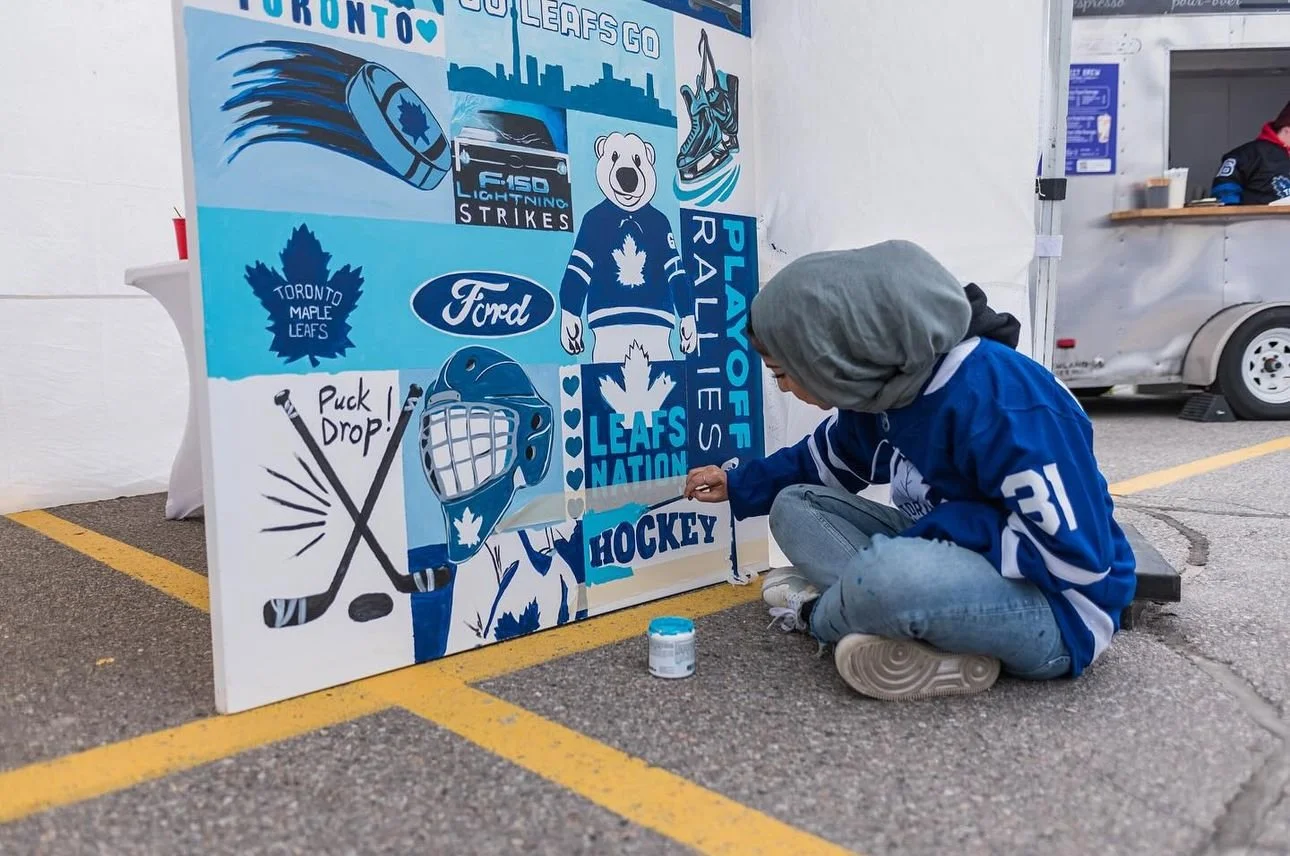 A person sitting on the ground painting a large Toronto Maple Leafs-themed mural with blue and white hockey-related images and text, including a hockey player, blue maple leaf, and the words "Leafs Nation" and "Hockey."