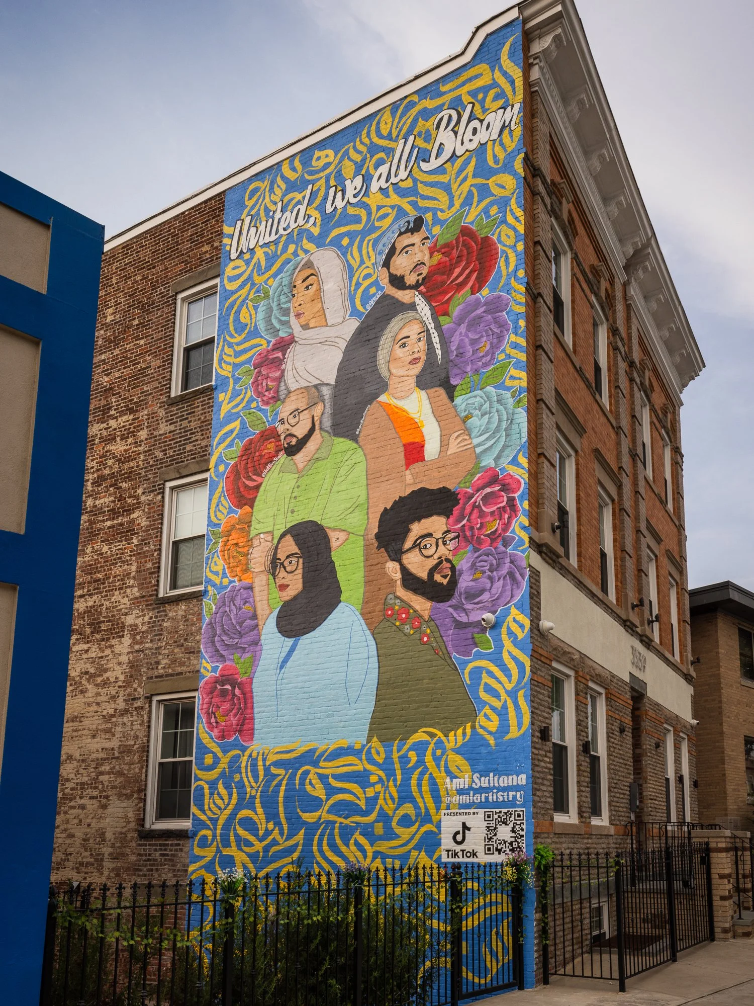 Colorful mural on the side of a brick building featuring illustrated portraits of diverse people surrounded by flowers and a yellow and blue decorative background. The text reads 'United, we all Bloom' and includes a QR code and TikTok logo.