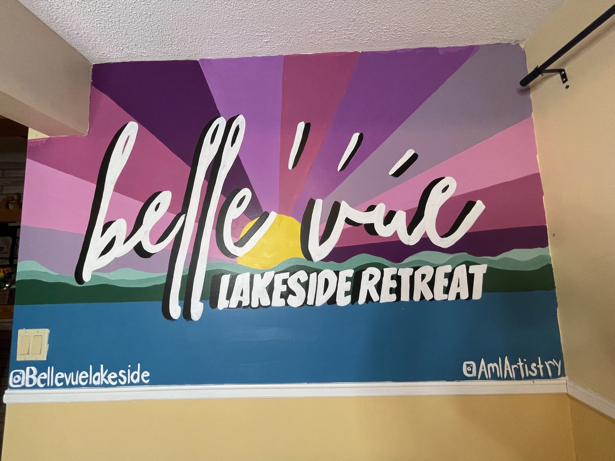 Colorful mural with the text 'Hello Wall Lakeside Retreat' over a landscape with a setting sun, mountains, and water. Social media handles '@Bellevuelakeside' and '@AmlArtistry' are at the bottom.
