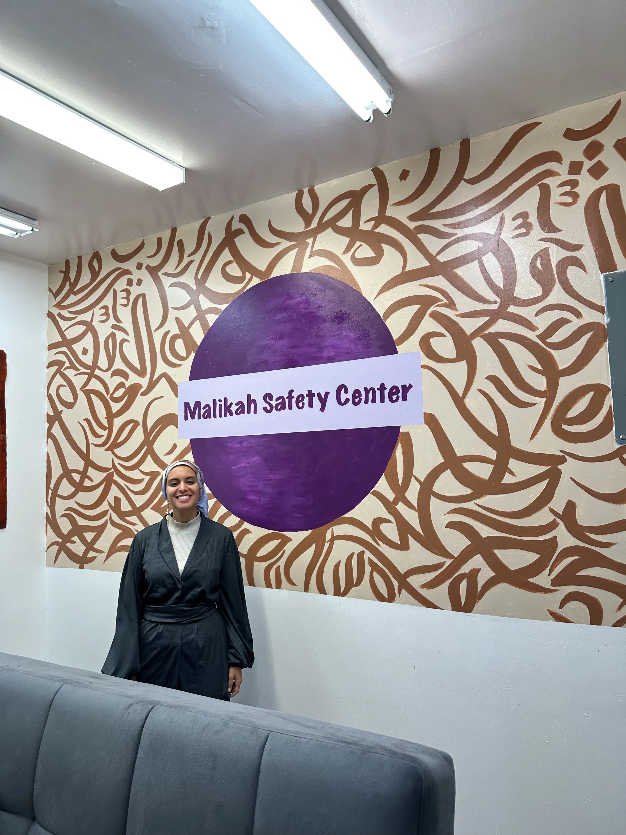 A woman standing in front of a mural with the text 'Malikah Safety Center' inside a purple circle.