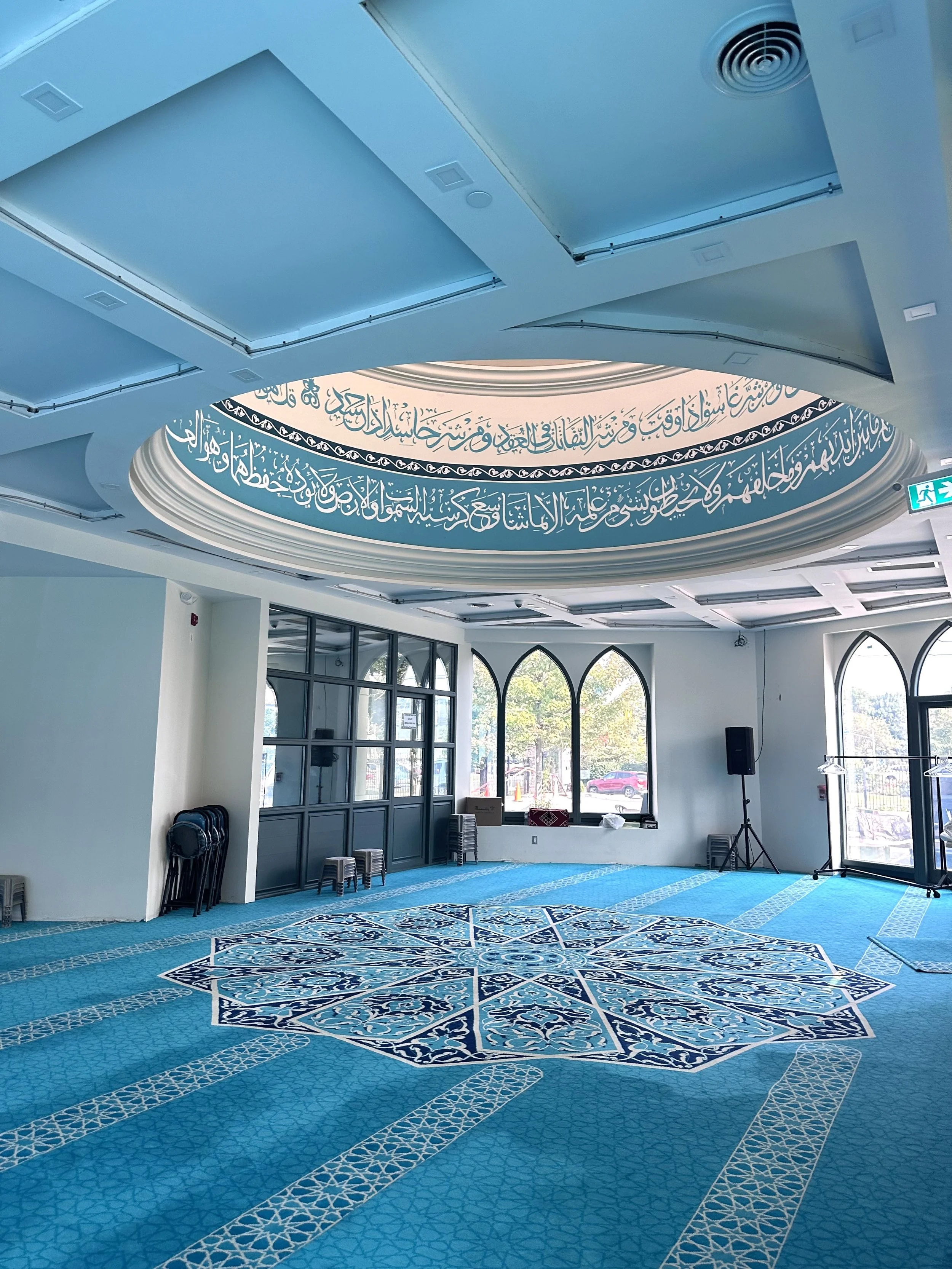 Interior of a mosque prayer room with a blue carpet, arched windows, and Arabic calligraphy on the ceiling